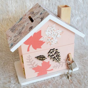 May include: A light pink wooden house-shaped piggy bank with a floral patterned roof and a chimney. The exterior features decorative cutouts of maple and oak leaves in shades of coral and white. A silver padlock with a key is attached to the side.