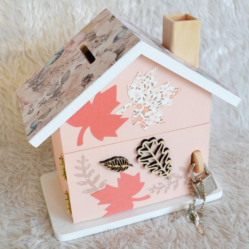 May include: A light pink wooden house-shaped piggy bank with a floral patterned roof and a chimney. The exterior features decorative cutouts of maple and oak leaves in shades of coral and white. A silver padlock with a key is attached to the side.
