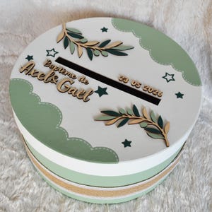 Peut inclure: Une boîte à souvenirs ronde, blanche et verte, avec une fente sur le dessus. Elle est décorée de branches d'olivier en bois, d'étoiles, et des inscriptions "Baptême de Axel & Gael" et la date "23 05 2023". Un ruban orne le milieu de la boîte.