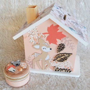 Peut inclure: Une tirelire décorative en forme de maison en bois avec une façade pêche, représentant un cerf, des motifs floraux et une grande feuille corail. Une petite boîte ronde avec un design de souris et de dent est à côté.