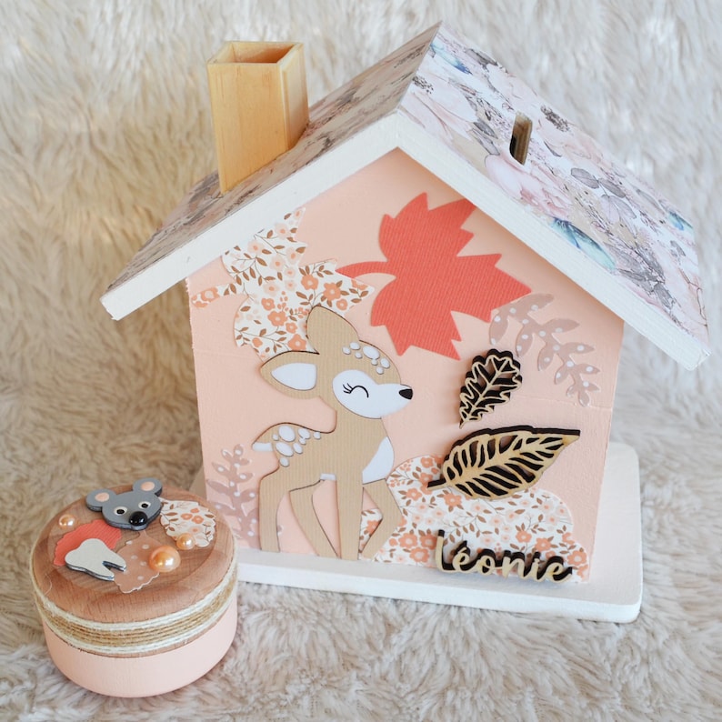 May include: A wooden house-shaped piggy bank painted in peach with a floral roof, featuring a deer cutout, autumn leaves, and the name L&eacute;onie. Beside it sits a small round wooden box decorated with a mouse, a tooth cutout, and peach beads.