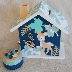 May include: A house-shaped wooden coin bank with a dark blue front, a light green leaf-patterned roof, and a tan chimney. The front features a tan deer cutout, gold glitter leaves, and dark leaf accents. Beside it sits a small round wooden box.
