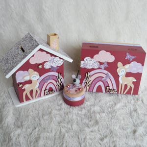 May include: A wooden house-shaped piggy bank and a rectangular wooden box, both painted maroon with a fawn, rainbow, clouds, and butterflies. A small round container with a mouse topper sits between them. Both items feature the name Eva.