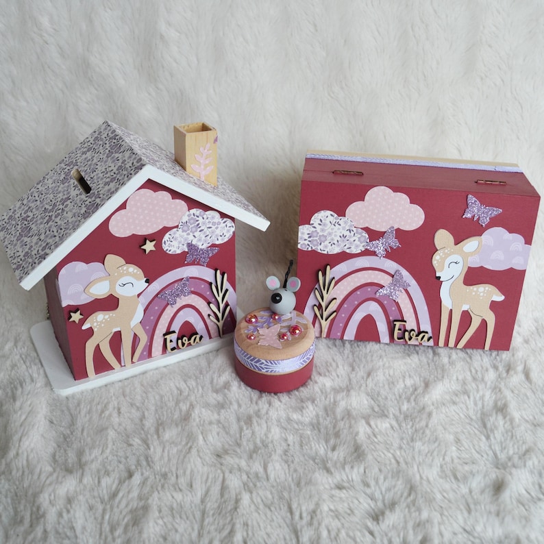 May include: A wooden house-shaped piggy bank and a rectangular wooden box, both painted maroon with a fawn, rainbow, clouds, and butterflies. A small round container with a mouse topper sits between them. Both items feature the name Eva.