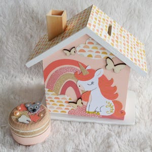 Peut inclure: Une tirelire en bois en forme de maison avec une façade rose, ornée d'une licorne blanche à corne dorée, d'un arc-en-ciel pailleté et de papillons en bois. À côté se trouve une petite boîte ronde en bois décorée d'une souris, d'étoiles et de perles.