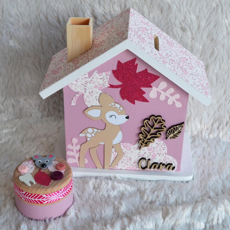 May include: A pink wooden house-shaped piggy bank with a floral roof, a chimney, and a cutout for coins. The front features a deer illustration, glittery red leaves, and the name Clara. Beside it sits a small round pink container with a mouse topper.
