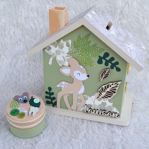 May include: A wooden house-shaped piggy bank painted sage green with a fawn illustration, leaf cutouts, and the name Louison in script. Beside it sits a small round wooden box decorated with a mouse face, buttons, and a tooth-shaped cutout.