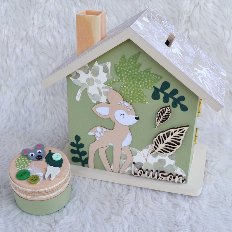 May include: A wooden house-shaped piggy bank painted sage green with a fawn illustration, leaf cutouts, and the name Louison in script. Beside it sits a small round wooden box decorated with a mouse face, buttons, and a tooth-shaped cutout.