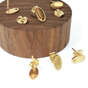 Op de afbeelding: Vijf gouden ovale oorbelblanken met een pen en een gat in het midden. De oorbelblanken worden gepresenteerd op een houten oppervlak.