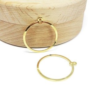 May include: Two gold-coloured, circular earring components with a small loop at the top, resting on a white surface. A light-coloured wooden box is in the background. The earring components are simple and elegant.
