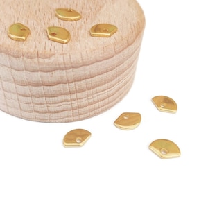 Peut inclure: Petits charms métalliques dorés, de forme plate en éventail, avec un petit trou en haut. Plusieurs breloques sont disposées sur une surface en bois clair, avec quelques autres éparpillées.