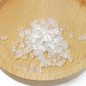 Puede incluir: Un conjunto de topes de pendientes transparentes, dispersos sobre una bandeja de madera marrón claro. Los topes son pequeños, translúcidos y tienen forma redondeada con un agujero para el poste. La bandeja es circular con un borde elevado.