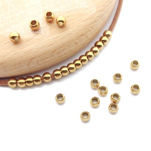 May include: Gold-toned metal beads for jewelry making. The beads are round and have a smooth finish. There are several beads scattered on a white surface, and a strand of beads is strung on a brown leather cord.