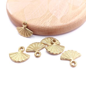 Puede incluir: Seis abalorios de metal de color dorado en forma de abanico con un bucle en la parte superior. Los abalorios están dispuestos sobre una superficie de madera clara.