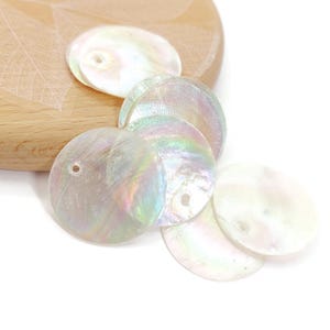 May include: Iridescent, round mother-of-pearl shell beads with a hole for stringing. The beads are a mix of white, cream, and pastel colors. They are scattered on a light-colored wooden surface.