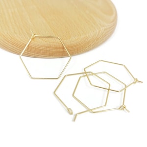 Pode incluir: Três brincos de argola hexagonais de ouro. Os brincos são feitos de fio fino e têm um diâmetro de aproximadamente 5 cm.