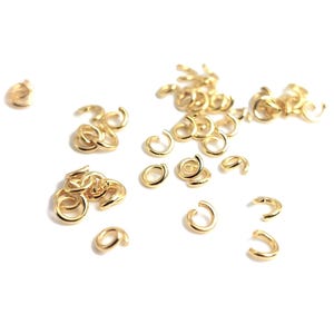 Op de afbeelding: Een verzameling kleine, goudkleurige sprongringen verspreid op een wit oppervlak. Deze open ringen worden gebruikt voor het maken van sieraden en knutselen. De ringen zijn in verschillende maten en vormen, met een glanzende, metalen afwerking.