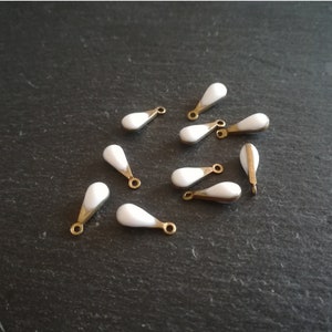 Könnte beinhalten: Zehn weiße tropfenförmige Anhänger mit goldfarbenen Metallscharnieren. Die Anhänger sind auf einer dunkelgrauen Oberfläche verstreut.