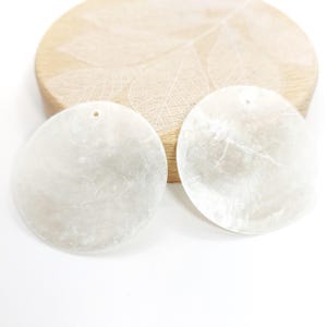 Könnte beinhalten: Zwei runde, irisierende weiße Muschelanhänger mit einem kleinen Loch zum Auffädeln. Die Anhänger sind vor einem hellen Holzgrund mit einem Blattmuster platziert.