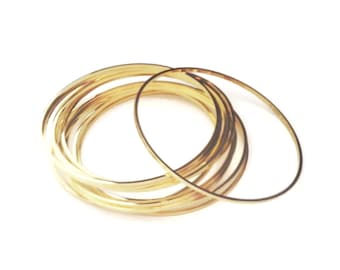 May include: A collection of thin, gold-colored bangles, some overlapping, against a white background. The bangles have a smooth, reflective surface, and the light creates highlights and shadows, giving them a three-dimensional appearance.