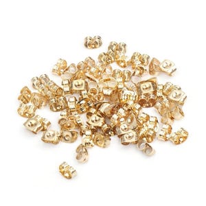 May include: A large pile of gold-colored earring backs. The backs are made of metal and have a small loop on the top. They are designed to be used with post earrings.