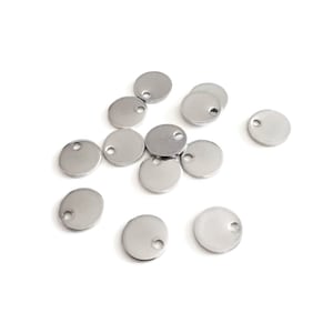 Op de afbeelding: Een groep ronde zilveren metalen hangers met een klein gaatje in het midden. De hangers liggen verspreid op een wit oppervlak.