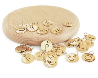 May include: A collection of gold-toned metal charms with different letters of the alphabet engraved on them. The charms are scattered on a wooden surface.