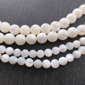 Può includere: Una collana di perle bianche di madreperla. Le perle sono rotonde e hanno una finitura liscia e lucida. Le perle sono infilate su un filo trasparente.
