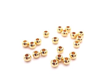 May include: A collection of small, shiny, gold-colored beads scattered on a white surface. Each bead has a rounded shape with a hole running through the center. These beads are likely used for jewelry making or other craft projects.