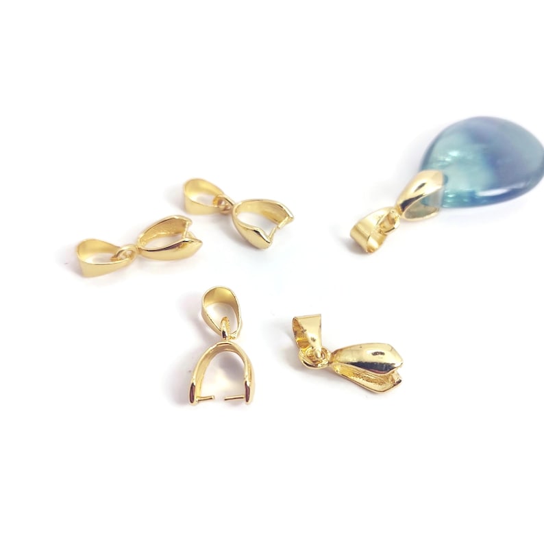 May include: A collection of gold-coloured bail findings, used for attaching pendants to necklaces or bracelets, are scattered on a white surface. One finding is attached to a blue and green stone.