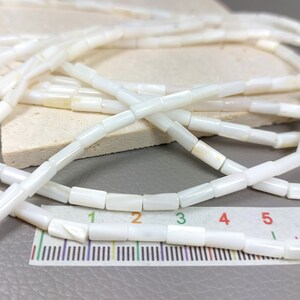 May include: A strand of white shell beads, each bead is a rectangular tube shape. The beads are laid out on a ruler, showing that each bead is approximately 1/2 inch long.