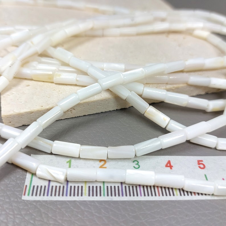 May include: A strand of white shell beads, each bead is a rectangular tube shape. The beads are laid out on a ruler, showing that each bead is approximately 1/2 inch long.