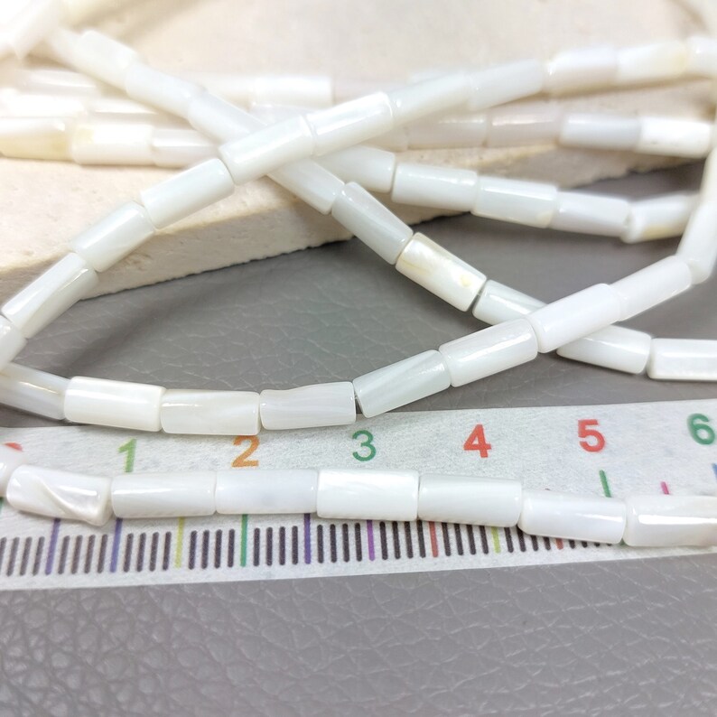 May include: A strand of white mother of pearl beads, each bead is a rectangular shape. The beads are laid out on a ruler, showing the size of the beads. The beads are approximately 1/2 inch long.