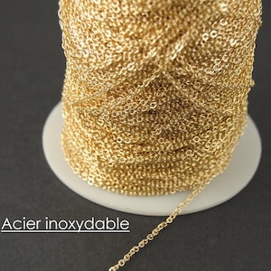 Puede incluir: Un carrete de cadena dorada, con un diseño delicado y enlazado. La cadena está enrollada en un carrete de plástico blanco. El texto "Acier inoxydable" es visible en la esquina inferior izquierda.