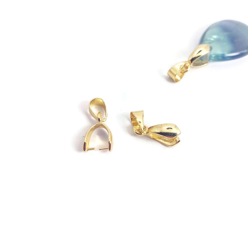 May include: Multiple gold-coloured pendant bails are shown against a white backdrop. The bails are in different positions, highlighting their design. One bail is attached to a blue and clear stone. These findings are used for jewellery making.