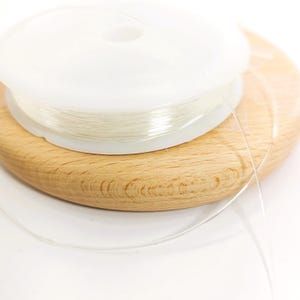 Peut inclure: Une bobine de fil élastique transparent pour perles est posée sur une base en bois clair. Le fil est enroulé sur une bobine en plastique blanc. Une longueur de fil s'étend, suggérant son utilisation dans la création de bijoux ou l'artisanat.