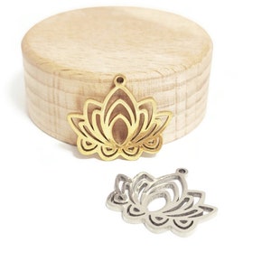 Peut inclure: Deux pendentifs en métal en forme de fleur de lotus, l'un doré et l'autre argenté, présentés sur un bloc de bois clair. Les pendentifs ont un design ajouré détaillé. Le fond est blanc.