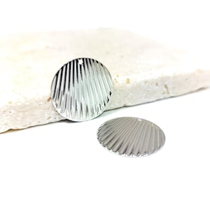 Puede incluir: Dos abalorios de metal de color plateado en forma de concha con textura estriada. Los abalorios tienen un agujero en la parte superior para sujetarlos a joyas u otras manualidades.