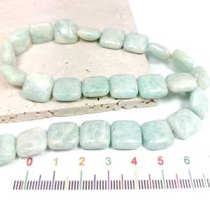 Peut inclure: Des rangées de perles de pierres précieuses Amazonite vert pâle de forme carrée. Les perles ont une qualité légèrement translucide et une finition lisse et polie. Les perles sont présentées sur une surface de couleur neutre avec une règle pour la référence de taille.
