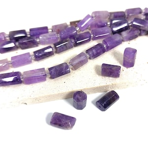 May include: A strand of purple amethyst gemstone beads, with a faceted, rectangular shape. The beads are strung on a clear thread and are arranged in a loose, flowing pattern. There are also several loose beads scattered around the strand.