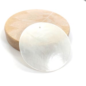 Può includere: Un ciondolo rotondo bianco perlaceo con un piccolo foro nella parte superiore, appoggiato su una base di legno circolare marrone chiaro. La base in legno presenta un motivo a foglie. Il ciondolo ha una superficie liscia e riflettente.