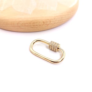 Peut inclure: Un fermoir mousqueton ovale doré avec un centre orné de cristaux étincelants. Le fermoir est posé sur un fond blanc, avec une partie d'une surface en bois à motif de feuilles visible dans le coin supérieur gauche. Idéal pour la création de bijoux.