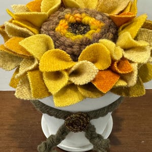 May include: A handcrafted felt sunflower arrangement in a white pot. The sunflower features yellow, orange, and tan petals surrounding a brown center. The pot is adorned with a green rope and a decorative bronze-colored embellishment.