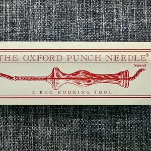 May include: A close-up of a packaging box for The Oxford Punch Needle, a rug hooking tool. The box is white with red text and a red illustration of the tool. The text on the box reads "The Oxford Punch Needle" and "A Rug Hooking Tool". The tool is labeled "Natural".