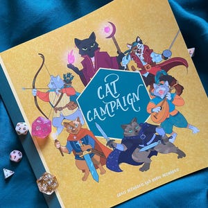 May include: A yellow and green book titled "Cat Campaign" with illustrations of cats in fantasy attire. Several dice are scattered around the book. The book's cover features a teal hexagon with the title in white script.