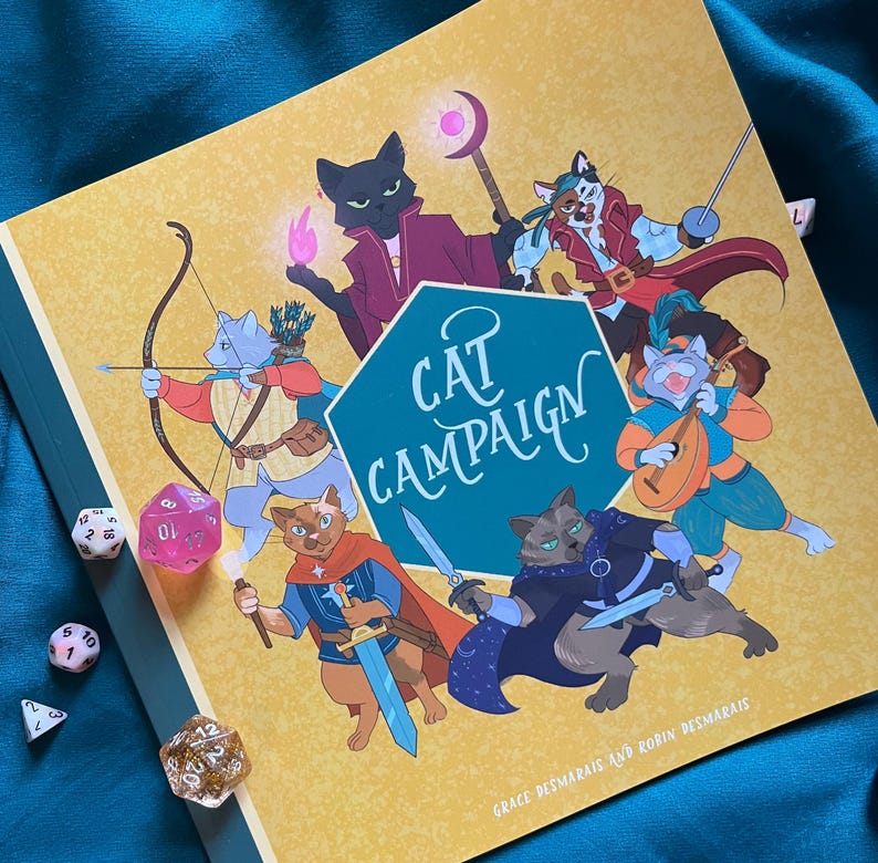 May include: A yellow and green book titled "Cat Campaign" with illustrations of cats in fantasy attire. Several dice are scattered around the book. The book's cover features a teal hexagon with the title in white script.
