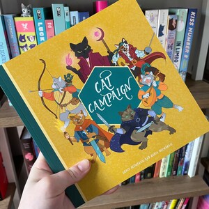 May include: A hardcover book titled "Cat Campaign" with a mustard yellow cover and a green spine. The cover features cartoon cats in various fantasy roles. The book is held in a person's hand, with a bookshelf in the background.