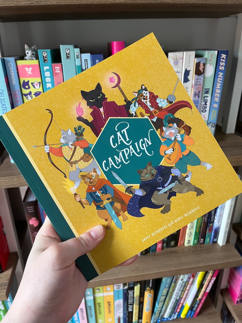 May include: A hardcover book titled "Cat Campaign" with a mustard yellow cover and a green spine. The cover features cartoon cats in various fantasy roles. The book is held in a person's hand, with a bookshelf in the background.
