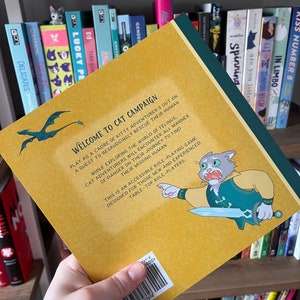 May include: The back cover of a book titled "Welcome to Cat Campaign" is held up, revealing a cartoon cat character with a sword. The cover is yellow with green and blue accents. The book is a tabletop role-playing game.