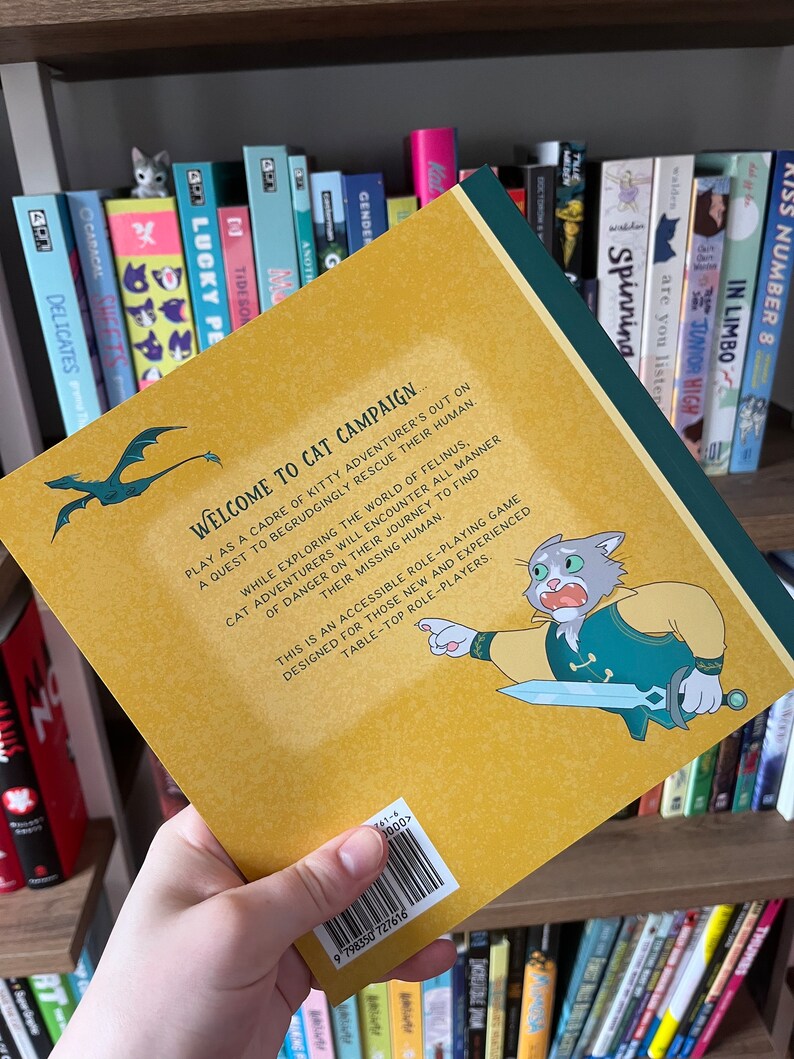 May include: The back cover of a book titled "Welcome to Cat Campaign" is held up, revealing a cartoon cat character with a sword. The cover is yellow with green and blue accents. The book is a tabletop role-playing game.